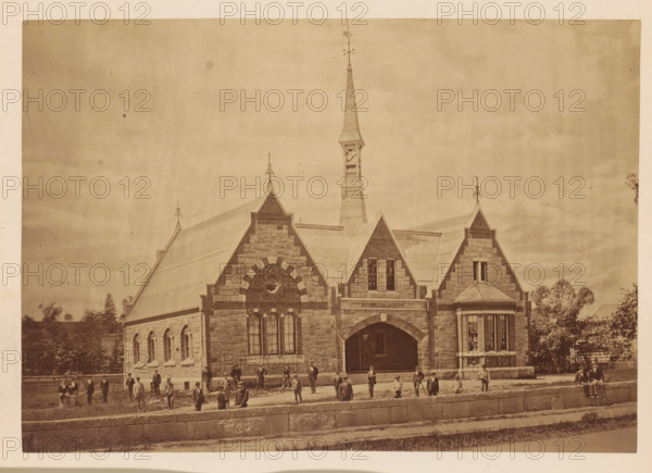 Memorial of William R. Dimmock, master of Adam Academy, photograph, page, Quincy, c. 1873 - in or before 1878, photographer, North America, Massachusetts, paper, albumen print, height 128 mm x width 182 mm, learned society, academy of sciences, anonymous historical persons depicted in a group, in a group-portrait, façade (of house or building), A stone building with steep gabled roofs and a tall central spire topped with a weather vane fills the scene, showing arched windows and a large arched entrance beneath decorative stonework; people stand and sit on the lawn and along a low stone wall in front of the building, and trees and a distant fence are visible at the sides.
