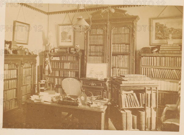 Memorial of William R. Dimmock, master of Adam Academy, photograph, page, Quincy, c. 1873 - in or before 1878, photographer, North America, Massachusetts, paper, albumen print, height 126 mm x width 178 mm, learned society, academy of sciences, anonymous historical persons depicted in a group, in a group-portrait, façade (of house or building), A sepia-toned room with tall wooden bookshelves filled with books, some with glass-fronted cabinets and ornate carved tops; a large central cabinet with double doors and shelves of books stands against the back wall, flanked by framed pictures and a bust on a shelf; a desk in the foreground cluttered with papers, small objects, and a tabletop clock, with a cane-backed chair pushed close to it; stacks of large books and rolled materials sit on a smaller table and open shelving beside the desk; a pendant light hangs from the ceiling above the central area and decorative trim runs along the top of the walls; various framed artworks hang on the wall...