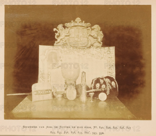 Collection of souvenirs of Admiral Michiel de Ruyter and his son, Souvenirs van Adm. de Ruyter en zijn zoon, photograph, page, number: 'No. 840, 842, 843, 846, 847, 853, 855, 856, 859, 860*, 945, 952.', photographer: Pieter Oosterhuis, Amsterdam, 1876, paper, albumen print, albumendruk, height 127 mm x width 162 mm, glass, rummer, container of precious metal, medal, badge of a guild, Historische Tentoonstelling Amsterdam, Oudemannenhuis (Amsterdam), Period, A sepia-toned photograph shows a tabletop display of objects arranged in front of an ornate backdrop topped by a decorative crest flanked by a lion and a horse. In front of the backdrop a large goblet-like vessel sits centrally, accompanied by a tall narrow spout vessel and a smaller rounded vessel nearby. To the right of the vessels sits a dark fabric cap or cushion decorated with light floral embroidery and trim, with a cylindrical rod resting beside it. To the left a rectangular block with an inscribed band is visible next to a s...