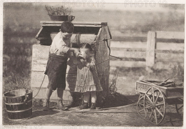 Boy lets a little girl drink water at a well, page, photomechanical print, photographer: Geo B. Wood, North and Central America (possibly), cliché maker: The Photo-gravure Co. (possibly), New York (city), c. 1884 - in or before 1889, printer (person), North America, paper, photoaquatint, height 118 mm x width 174 mm, boy (child between toddler and youth), well, toddler (female), A sepia-toned scene shows two children standing beside a weathered wooden well structure; an older child in rolled-sleeve shirt and dark knee-length trousers leans over and holds a container to the face of a younger child in a patterned dress, while the younger child lifts hands toward the container. Nearby are a wooden bucket with metal bands, a small wooden cart with spoked wheels containing flat objects, and a shallow round basin on the ground. The well structure has coiled rope and a potted plant placed on its top, and a simple horizontal fence runs in the blurred background.