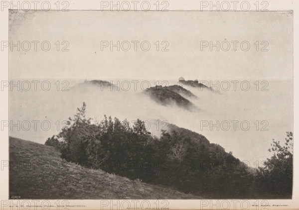 Mount Hamilton, View of Mount Hamilton and the Lick Observatory, page, photomechanical print, inscription: 'Kurtz', photographer: Sherburne Wesley Burnham, Hamilton, Mount, cliché maker: Kurtz, 1887 - 1907, North America, California, Santa Clara (California), paper, autotype, height 115 mm x width 172 mm, mist, fog, mountains, meteorological observatory, Lick Observatory, A grayscale landscape photograph showing a hillside in the foreground with grasses and clustered trees and shrubs, a series of rounded ridges and peaks rising and fading into a low-lying layer of mist that fills the middle distance, and a faint sky above, all framed by a narrow border around the image.