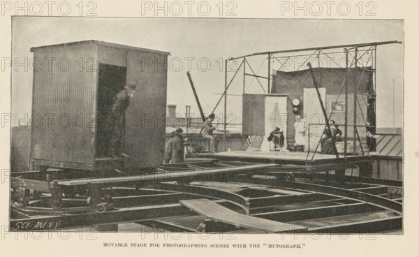 Movable Stage for Photographing Scenes with the 'Mutograph', Page, photomechanical print, inscription: 'SCI. AM. NY', maker: unknown, c. 1892 - in or before 1897, paper, autotypie, height 104 mm x width 191 mm, the stage and its equipment, A large outdoor scene on a raised platform shows a corrugated rectangular structure with an open doorway on the left and a scaffolding-like frame on the right supporting a curtained backdrop and a small enclosed area. Several people are present: one person leaning out from the doorway of the corrugated structure, a few individuals standing or seated on the platform near the frame, and two seated figures inside the framed area near the backdrop. The platform rests on visible metal beams and rails with wheels and mechanical elements beneath and around it. A plank crosses part of the platform in the foreground, and a tall angled pole or boom rises from near the center toward the right. The overall image has a grainy, monochrome appearance.