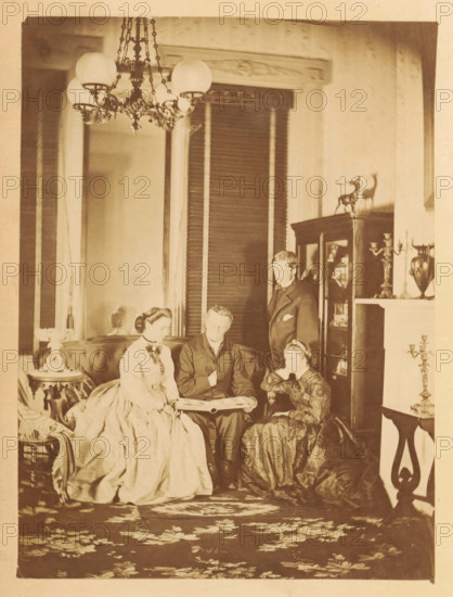 Two men and two women look at a photo album in a living room, photograph, page, photographer: JC Browne, c. 1861 - in or before 1866, paper, albumen print, albumendruk, height 167 mm x width 125 mm, achter semi-transparent cover., adult woman, adult man, living-room, parlour, sitting-room, A sepia-toned interior scene showing four people arranged around a low table, with two women seated on a curved upholstered sofa, a man seated between them holding open a large book or album, and another man standing behind them near a glass-fronted cabinet; the seated women wear full, floor-length dresses with fitted bodices and the men wear dark suits; the room includes a patterned rug, ornate chandelier with globe shades hanging from the ceiling, decorative wood trim and tall shutters behind the group, a display cabinet topped with small stag figurines, a mantel with candelabra and a vase, a small round side table with a decorative lamp, and additional furniture and ornamentation visible around th...