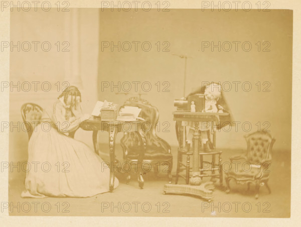 A little boy plays at a table, while a girl reads at another table, photograph, page, photographer: James Inglis, c. 1861 - in or before 1866, paper, albumen print, height 115 mm x width 157 mm, behind semi-transparent cover., girl (child between toddler and youth), toddler (male), A sepia-toned indoor scene showing a woman seated and leaning her head on her hand next to an ornate round wooden table with papers and a decorative box on it, upholstered chairs around the table, a small wooden stand holding a camera and a doll positioned behind the camera, another small upholstered chair to the right with a doll sitting on it, and various small objects and fabric on the floor in front of the stand, all set against a plain wall.