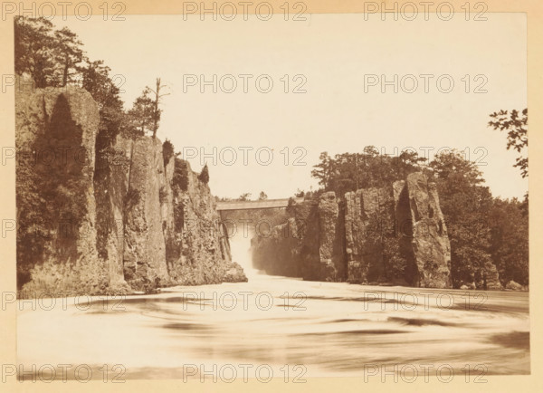 View of the Great Falls of the Passaic River, New Jersey, photograph, page, photographer: J. Reid, Passaic, c. 1861 - in or before 1866, North America, paper, albumen print, height 117 mm x width 166 mm, behind semi-transparent cover., waterfall, A sepia-toned landscape showing a river flowing between tall vertical rock cliffs, with water appearing smooth and blurred in the foreground, rocky faces rising steeply on both sides, trees and vegetation atop and along the cliffs, and a narrow bridge spanning the gap near the upper center of the scene.