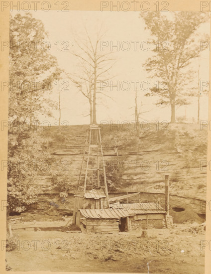 View of an oil well, photograph, page, photographer: Rogers, SG & TW, North and Central America (possibly), c. 1861 - in or before 1866, paper, albumen print, height 156 mm x width 119 mm, behind semi-transparent cover, pumping of oil, A sepia-toned photograph showing a weathered wooden structure with a slanted corrugated roof and rough plank walls situated at the base of a rocky cliff, a tall wooden framework or derrick rising from the structure with visible crossbeams and supports, a narrow deck or platform extending alongside the building, a vertical metal pipe or smokestack near the right side of the structure, layered rock faces forming the cliff behind with small ledges and vegetation, trees lining the top of the cliff including a mix of leafed and bare-branched trunks, dense foliage visible along the left edge, and a grassy foreground with a small stump and scattered ground cover.
