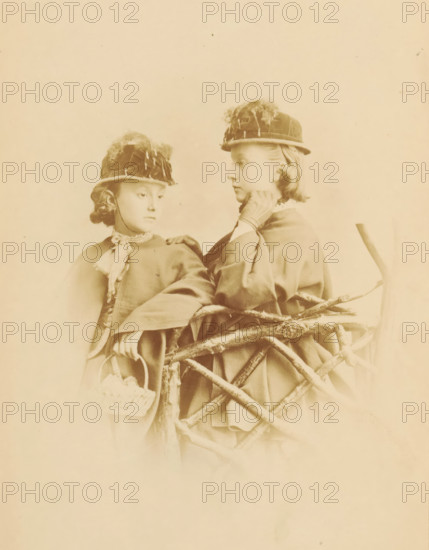 Portrait of two unknown young girls with hats, photograph, page, photographer: William Notman, c. 1861 - in or before 1866, paper, albumen print, height 157 mm x width 115 mm, behind semi-transparent cover., historical persons not known by name - BB - woman, girl (child between toddler and youth), Two young girls wearing round hats with decorative feathers stand behind a rustic wooden fence made of intertwined branches, both dressed in cloaks with high collars and one girl holding a small woven basket; one girl looks slightly downward while the other faces her with a hand near her neck, and the scene is presented in a soft sepia tone.