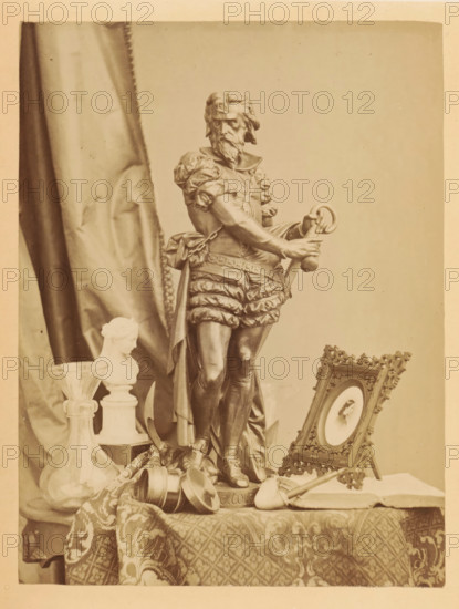 Bronze sculpture, photo frame, pipe and other items on a table, photograph, page, photographer: Frederick A. Wenderoth, c. 1861 - in or before 1866, paper, albumen print, height 166 mm x width 125 mm, behind semi-transparent cover., sculpture, pipe, tobacco, A photograph of a tabletop still life dominated by a tall statue of a bearded man in ornate period clothing holding a ring and sword, standing on a pedestal, with a draped patterned cloth beneath; to the left of the statue there is a glass vessel with a wide base and a smaller bust on a plinth, and a tall folded fabric backdrop; to the right of the statue an ornate framed oval portrait rests on a small easel, and a smoking pipe lies on the table nearby; scattered around the base of the statue are small metal cups and a rolled textile, and decorative elements and fabric textures fill the foreground.