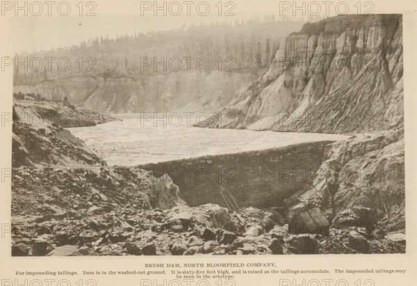View of Brush Dam in North Bloomfield, California, Brush Dam, North Bloomfield Company, page, photomechanical print, inscription: 'For impounding tailings. Dam is in the washed-out ground. It is sixty-five feet high, and is raised as the tailings accumulate. The impounded tailings may, be seen in the artotype.', North Bloomfield, c. 1885 - in or before 1890, North America, paper, collotype, photoprint, height 110 mm x width 180 mm, mine, A sepia-toned landscape photograph showing a wide river or reservoir stretching between steep, rocky banks with layered cliffs on one side and a rugged, rubble-strewn foreground on the other, the shoreline appearing uneven and eroded, with a high rim of cliffs displaying vertical striations and sloping faces, a treeline visible along the far ridge above the cliffs, scattered rocks and boulders in the foreground, and a generally overcast or hazy sky.