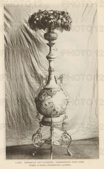 Lamp. American Art Fayence. Greenpoint, New York, Decorated lamp, page, photomechanical print, inscription: 'Height, 57 inches, circumference, 35 inches.', c. 1885 - in or before 1890, North America, paper, collotype, photoprint, height 183 mm x width 110 mm, lighting, lamps, An ornate floor lamp with a decorative base and stand is photographed against a wrinkled fabric backdrop, showing a detailed, rounded central body with floral reliefs and curved handles, a tall neck with carved sections and ornamental knobs, a wide fringed shade topped with clustered floral elements, and an elaborate tripod-like metal support with scrollwork feet.