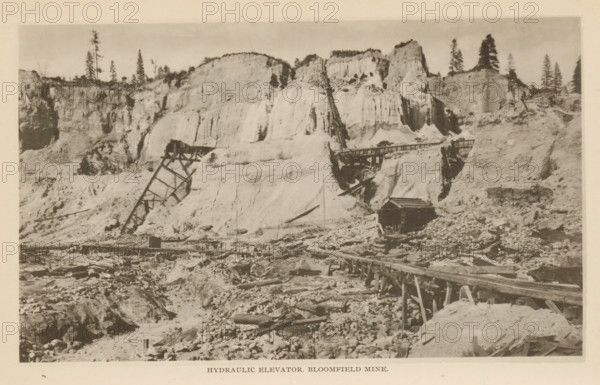 Hydraulic Elevator, Bloomfield Mine, View of a hydraulic elevator in the Bloomfield Mine, California, page, photomechanical print, North Bloomfield, c. 1885 - in or before 1890, North America, paper, collotype, photoprint, height 110 mm x width 183 mm, mine, A sepia-toned photograph of a rugged landscape with steep, light-colored cliff faces rising across the background, punctuated by scattered tall coniferous trees along the ridge. In front of the cliffs are remnants of wooden industrial structures, including angled supports and a long elevated timber chute extending up the slope and across a gap, with a small dark wooden building situated near the base of the cliffs. The foreground shows rough, rocky terrain strewn with debris and timber, and a raised wooden track or walkway running horizontally toward the right side of the image. The scene has a weathered, aged appearance.