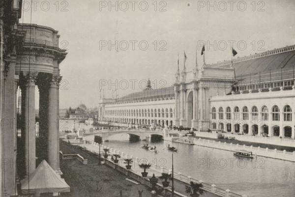 Manufacturer's buildings, world's fair, Chicago, page, photomechanical print, photographer: Charles Dudley Arnold, cliché maker: Benedict & Co., Geo. H., 1893, North America, Illinois, paper, autotypie, height 116 mm x width 173 mm, world fair, world exhibition, A riverside scene showing a large classical building with arched windows and tall decorative columns, a grand entrance with ornate towers and flags on top, and a long roof with a row of railings; in front of the building a curved bridge spans the water, several small rowboats and a larger boat float on the river, and promenades with lamp posts and potted plants run along the waterfront; on the left foreground a portion of another columned structure is visible with a tented canopy below and sculptural details on the column capitals, while distant buildings and trees appear along the far bank beneath an open sky.