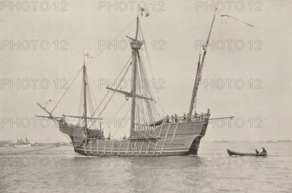 The 'Santa Maria', page, photomechanical print, photographer: J. Byron, cliché maker: Andrews & Son Co., J., c. 1888 - in or before 1893, paper, autotypie, height 108 mm x width 163 mm, ships (in general), A large wooden sailing ship with tall masts and rigging floats on calm water, its hull showing visible planking and a raised stern with a railing where several small figures stand, a smaller boat with two seated figures is alongside, flags fly from the mastheads and spars, rigging lines run between masts and down to the deck, and distant low shoreline and additional ships are visible on the horizon under a pale sky.