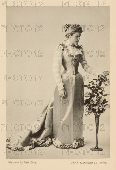 The infanta Eulalia, page, photomechanical print, photographer: GW Pach, cliché maker: Gutekunst & Co., F., Philadelphia, c. 1888 - in or before 1893, North America, paper, collotype, photoprint, height 155 mm x width 106 mm, Person, Eulalia van Bourbon, ruler, sovereign - BB - female ruler, A sepia-toned photograph shows a woman standing in profile with her head slightly turned downward toward a tall slender vase of flowers beside her. She is wearing a long, satin dress with a fitted bodice, puffed sleeves with horizontal bands, and a high collar adorned with a small decoration; the skirt extends into a ruffled train on the floor. Her hair is styled up and held with a ribbon or fabric accessory. One of her hands rests at her side while the other reaches toward a rose in the vase, which contains a stem with leaves and additional blooms and foliage. The background is plain and light, and the image has a narrow border around it.