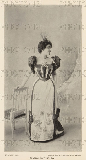 Flash-light study, page, photomechanical print, photographer: Charles Louis Klary, Paris, cliché maker: anonymous, c. 1890 - in or before 1895, paper, autotypie, height 192 mm x width 101 mm, adult woman, studio, A sepia-toned portrait of a woman standing next to a wooden chair, wearing a floor-length dress with a fitted, striped bodice and puffed sleeves, a contrasting apron-like front panel adorned with floral embellishments, and large bows at the hips; she has short curly hair topped with a small feathered headpiece and wears a necklace, her left hand resting on the back of the chair while she looks to her right, and a patterned backdrop is visible behind her.