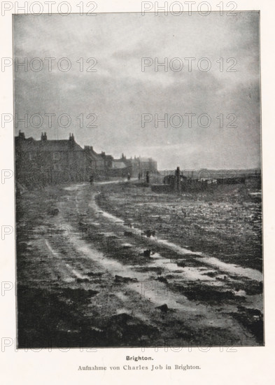 Brighton, View of the Brighton coast, page, photomechanical print, photographer: Charles Job, cliché maker: anonymous, printer (person): Wilhelm Knapp Verlag, Halle an der Saale, c. 1893 - in or before 1898, printer (person), Saxony-Anhalt, paper, autotypie, height 162 mm x width 120 mm, coast, A grainy black and white scene shows a wide muddy shoreline with visible wet ruts and patches of darker mud leading toward the horizon, a row of low buildings with pitched roofs and chimneys lines the left side, several small human figures stand or walk near the distant buildings, a faint line of structures and a low pier or breakwater extend into the water on the right, and a broad overcast sky fills the upper portion of the image.