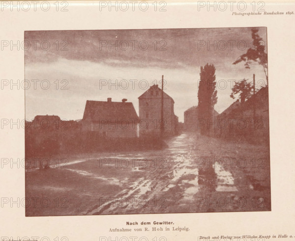 Nach dem gewitter, View of a village after bad weather, page, photomechanical print, photographer: R. Hoh, cliché maker: anonymous, printer (person): Wilhelm Knapp Verlag, Halle an der Saale, c. 1893 - in or before 1898, printer (person), Saxony-Anhalt, paper, autotypie, height 115 mm x width 152 mm, city-view, and landscape with man-made constructions, A sepia-toned image showing a street scene with dark silhouettes of buildings and tall narrow trees against a cloudy sky, a wet reflective road surface leading toward the foreground with visible puddles and shiny textures, rooftop shapes and chimneys outlined along the horizon, a taller building with a pitched roof near the center, and scattered vegetation and utility poles framing the scene.