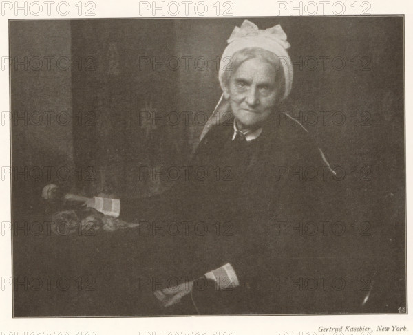 Portrait of an unknown old woman, page, photomechanical print, photographer: Gertrude Käsebier, cliché maker: Meisenbach, Riffarth & Cie. (possibly), Berlin, c. 1898 - in or before 1903, paper, autotypie, height 136 mm x width 179 mm, Print on fourth unnumbered page after page 182., historical persons not known by name - BB - woman, old woman, A monochrome photograph of an older woman seated against a dark background wearing a light headscarf tied in a bow and a dark garment with a visible collar, her face turned slightly toward the camera with a neutral expression, her hands resting in her lap with one hand holding a folded cloth, and a small table beside her holding a piece of folded fabric and some rounded objects.