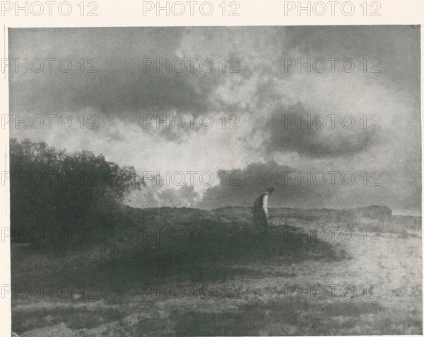 Man in a landscape, page, photomechanical print, photographer: Scholz, Th. & R., cliché maker: Meisenbach, Riffarth & Cie. (possibly), Berlin, c. 1898 - in or before 1903, paper, autotypie, height 139 mm x width 184 mm, Print on third unnumbered page after page 234., landscapes, adult man, A grainy black and white scene showing a wide sky filled with large, textured clouds above a dark landscape; near the horizon a solitary figure stands with a slightly bent posture wearing a darker outfit against a lighter background; to one side a dense cluster of foliage or trees creates a silhouette that contrasts with the open expanse, and the foreground shows uneven ground with varied tones and subtle texture.