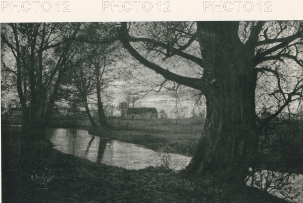 Landscape, with a house in the background, page, photomechanical print, photographer: Heinrich Bachmann, cliché maker: Meisenbach, Riffarth & Cie. (possibly), Berlin, 1903, paper, autotypie, height 134 mm x width 215 mm, Print on second unnumbered page after page 262., farm or solitary house in landscape, A wide dark-toned scene showing large tree trunks and bare branches in the foreground framing a calm reflective body of water that runs across the lower portion of the image, with the branches creating intricate silhouettes against a lighter sky; in the midground a simple rectangular building with a pitched roof sits on a flat area beyond the water, and sparse trees and low vegetation surround the building and shoreline, while the foreground ground appears uneven and textured.