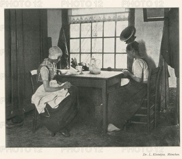 Two women sitting at a table doing needlework, page, photomechanical print, photographer: LL Kleintjes, cliché maker: anonymous, c. 1898 - in or before 1903, paper, autotypie, height 139 mm x width 174 mm, Print on second unnumbered page after page 290., adult woman, mending, repairing, clothes, A black and white scene of two seated women at a wooden table in front of a multi-paned window, one woman on the left wearing a head covering and an apron with hands in her lap, the other on the right leaning slightly forward with hands near the table edge; on the table there are a teapot, a cup, a plate, and some small items, a coat hangs on the wall beside the window, and light comes through the window panes into the room.