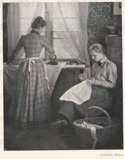 Woman ironing and a woman working with needles, page, photomechanical print, photographer: Albert Gottheil, cliché maker: Meisenbach, Riffarth & Cie. (possibly), Berlin, c. 1898 - in or before 1903, paper, autotypie, height 177 mm x width 144 mm, Print on second unnumbered page after page XI., sewing, ironing, A grayscale scene shows two women in a domestic interior near a window with sheer curtains; one woman stands with her back mostly to the viewer wearing a long plaid dress and a tied apron, her hair pulled up, and she has one hand on an iron placed on a covered table; the other woman sits to the right facing slightly downward, wearing a striped blouse and a dark skirt, holding a piece of cloth in her lap and appearing focused on it; a patterned wallpaper panel is visible behind the seated woman, a wooden cabinet edge appears nearby, and a woven basket with fabric and a long ribbon rests on the floor in the foreground.