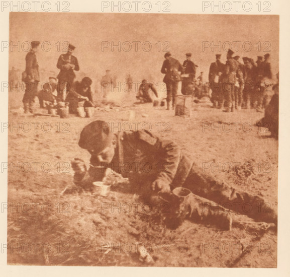 Soldier eating in a reclining position, page, photomechanical print, photographer: Th. Schneider, clichémaker: anonymous, c. 1898 - in or before 1903, paper, photoaquatint, height 146 mm x width 156 mm, Print two of fourteen prints at the back of the book., private soldier, eating, A grainy sepia-toned photograph showing a group of figures scattered across a flat, rough ground; in the foreground a person bent over with their head lowered and hands near a small bowl or dish, nearby containers and cloth lie on the ground; midground figures stand or crouch, some appearing to handle bowls or buckets while others stand upright wearing peaked caps and coats; a line of additional standing figures forms a loose grouping toward the background at the right edge, all rendered with soft, blurred outlines and muted tonal contrasts against a pale sky.