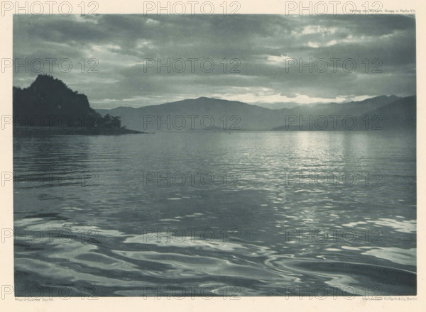 Am Luganer See, View of Lake Lugano, page, photomechanical print, photographer: Franz Goerke, Lugano, cliché maker: Meisenbach, Riffarth & Cie., Berlin, publisher: Wilhelm Knapp Verlag, Halle an der Saale, c. 1898 - in or before 1903, printer (person), Saxony-Anhalt, paper, photoaquatint, height 137 mm x width 195 mm, Print four of fourteen prints at the back the book., lake, mountains, A wide view of a calm body of water extending toward distant low mountains under a cloudy sky, with gentle ripples and reflections on the water surface, a dark silhouetted shoreline with a rocky promontory and some trees on the left, and layered mountain ridges fading into the background beneath scattered clouds.