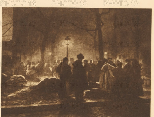 View of a market in Brussels, Sonnabendmarkt in Brüssel, page, photomechanical print, photographer: A. Cumont, Brussels, cliché maker: Büxenstein & Comp., Georg, Berlin, c. 1898 - in or before 1903, printer (person), paper, photoaquatint, height 134 mm x width 180 mm, Print five of fourteen prints at the back of the book., market, A dimly lit street scene at night showing a crowd of shadowy figures gathered under a tall lit lamppost, with silhouettes of people wearing coats and hats, dark tree trunks and branches rising behind them, areas of light and shadow creating a grainy, sepia-toned atmosphere, cobblestone pavement visible beneath the figures, and blurred building shapes and windows faintly visible in the background.