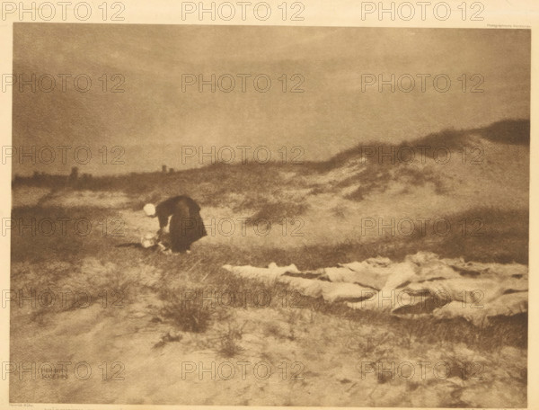 Woman washes clothes in the dunes, Wäscherin in der Düne, page, photomechanical print, photographer: Heinrich Kühn, cliché maker: Büxenstein & Comp., Georg, Berlin, c. 1898 - in or before 1903, printer (person), paper, photoaquatint, height 167 mm x width 227 mm, Print twelve of fourteen prints at the back of the book., adult woman, dunes (sea not visible), in the dunes, laundering, A grainy monochrome scene of a windswept landscape with low rolling dunes and rough grass, a solitary figure wearing a dark cloak bent forward near the ground and holding a pale round object, distant low horizon with indistinct vertical shapes, and textured foreground with scattered lighter patches on the sand.