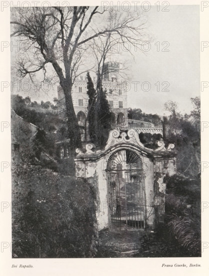 Bei Rapallo, View of a villa on a hill, near the town of Rapallo, page, photomechanical print, photographer: Franz Goerke, Rapallo, cliché maker: Meisenbach, Riffarth & Cie. (possibly), Berlin, c. 1898 - in or before 1903, paper, autotypie, height 199 mm x width 150 mm, Print on fourth unnumbered page after page 52., villa, gate, entrance, A black and white photograph showing a tall ornate stone gate with a decorative curved top and metal bars set in a weathered wall in the foreground, a narrow path leading up to the gate flanked by overgrown vegetation, a large bare-branched tree rising behind and to the left of the gate with additional leafless trees interwoven, and a multi-story building with arched windows and a crenellated tower partially visible through the trees in the midground, with distant foliage and a pale sky filling the background.