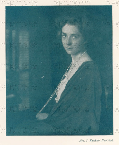 Portrait of an unknown woman with a long pearl necklace, page, photomechanical print, photographer: Gertrude Käsebier, cliché maker: Meisenbach, Riffarth & Cie. (possibly), Berlin, c. 1898 - in or before 1903, paper, autotypie, height 157 mm x width 130 mm, Print on second unnumbered page after page 80., historical persons not known by name - BB - woman, pearl, pearl necklace, A faded monochrome portrait of a woman shown from the torso upward, turned slightly to one side with her face looking toward the viewer, wearing a dark draped garment over a lighter blouse and a long beaded necklace that hangs down and is held in one hand, her hair styled away from her face, set against a dark, blurry background with a pale border surrounding the image.