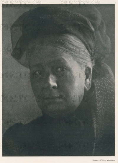 Portrait of an unknown woman, page, photomechanical print, photographer: Bruno Wiehr, cliché maker: anonymous, c. 1898 - in or before 1903, paper, autotypie, height 174 mm x width 129 mm, historical persons not known by name - BB - woman, A close-up black-and-white portrait of an older woman shown from the shoulders up, her face turned slightly toward the camera with a neutral expression, visible skin texture and fine hair pulled back under a dark head covering, one ear exposed, dark clothing with a textured fabric on one shoulder, and a plain darker background.