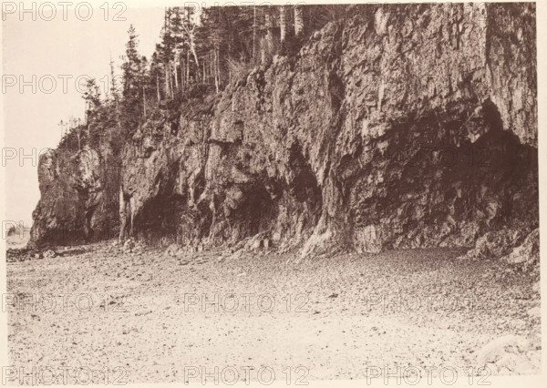 View of The Ovens rock formation on Mount Desert Island, Maine, The Ovens, Mount Desert, page, photomechanical print, c. 1875 - in or before 1880, North America, Mount Desert Island, paper, collotype, photoprint, height 131 mm x width 187 mm, rock formations, A wide sepia-toned photograph shows a rocky cliff face occupying the right and center of the image, with textured vertical rock strata, crevices, and shadowed recesses. The base of the cliff meets a pebbly or sandy shore that extends across the lower foreground. Along the top of the cliff there are slender tree trunks and sparse foliage, with some taller evergreens and thinner deciduous trunks visible against a pale sky. The left side of the image includes a smaller outcropping of rock with additional trees and a narrow strip of distant shoreline barely visible at the far left edge. The overall scene is detailed in high contrast tones and grainy texture.