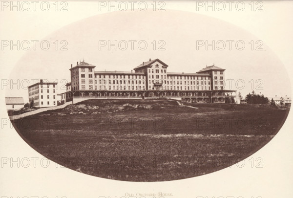View of Old Orchard House in Old Orchard Beach, Maine, Old Orchard House, page, photomechanical print, Old Orchard Beach, c. 1875 - in or before 1880, North America, paper, collotype, photoprint, height 191 mm x width 115 mm, hotel, hostelry, inn, A wide, oval-framed sepia photograph of a large multi-story building with a long covered porch running along its front, flanked by two taller corner sections with decorative rooflines and smaller attached wings; the building sits on a slightly raised grassy, rocky slope with a fenced area and a few smaller outbuildings and houses visible to the left and right, and a strip of open lawn filling the foreground.
