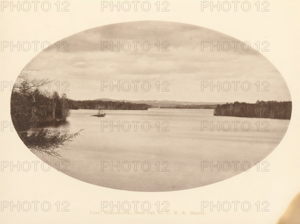 Lake Maranacook, from the MCRR Bridge, View of Lake Maranacook in Maine, page, photomechanical print, Winthrop, c. 1875 - in or before 1880, North America, Maine, paper, collotype, photoprint, height 115 mm x width 190 mm, river, An oval-shaped sepia photograph showing a wide calm lake with smooth water, a distant shoreline of trees and low hills, clusters of bare and leafed trees along the left and right edges, a small island with a few bare branches near the center, overcast sky with layered clouds, and tree branches extending into the frame from the lower left.