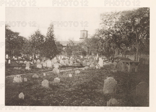 Cemetery of Christ Church in Cambridge, page, photomechanical print, photographer: William James Stillman, Cambridge (MA), cliché maker: anonymous, United States of America, c. 1866 - in or before 1876, North America, Massachusetts, paper, collotype, photoprint, height 128 mm x width 186 mm, church (exterior), churchyard (around church), A wide view of an outdoor scene showing an expanse of grass dotted with many upright stone markers of varying shapes and sizes, some rounded on top and others more irregular, arranged in rows and clusters across the foreground and middle ground, with a narrow path running among them; scattered trees of different heights stand throughout the area, their trunks and foliage creating shaded pockets, and larger trees form a dense line toward the background; beyond the trees a building with a square tower and a small roofed cupola is partially visible, its façade and roofline seen above a fence that runs along the rear of the field.