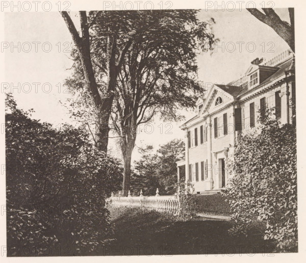 View of the house of Henry Wadsworth Longfellow in Cambridge, page, photomechanical print, photographer: William James Stillman, Cambridge (MA), cliché maker: anonymous, United States of America, c. 1866 - in or before 1876, North America, Massachusetts, paper, collotype, photoprint, height 148 mm x width 175 mm, housing, facade (of house or building), A sepia-toned scene showing the side of a large house with classical architectural details including shuttered windows, decorative cornices, and a triangular pediment with a rounded window, partially covered by climbing vines; in front of the house a low ornamental balustrade runs along a garden area, and tall trees with dense canopies rise beside and behind the house, their trunks and branches framing the view while hedges and shrubbery fill the foreground.