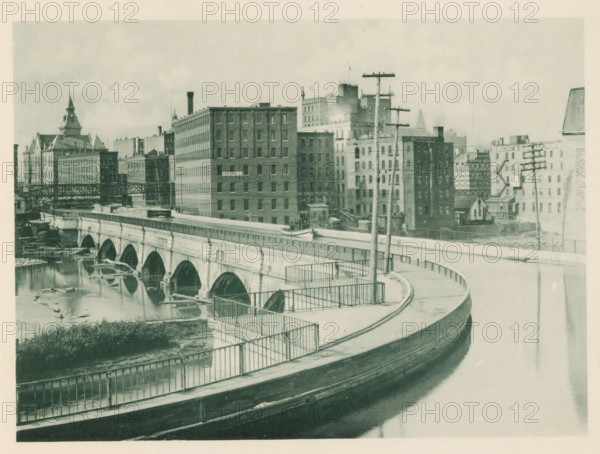 The Erie Canal viaduct at Rochester, NY, View of an aqueduct for the Erie over the Genesee in Rochester, page, photomechanical print, Rochester (New York), c. 1883 - in or before 1893, North America, paper, collotype, photoprint, height 140 mm x width 190 mm, canals, waters (in city), city-view, and landscape with man-made constructions, A curved roadway with metal railings runs along the foreground above a body of water, leading to a stone arched bridge that spans the river below; the bridge shows multiple rounded arches and reflections in the water. Beyond the bridge a cluster of multistory brick and stone buildings of varying heights lines the riverbank, with some rooftop structures, chimneys, and a tower with a pointed roof visible among them. Utility poles with crossarms and wires stand near the roadway and in front of the buildings. The riverbank shows exposed rocks and debris at the waterline, and a raised walkway or elevated structure extends across part of the scene. The photo...