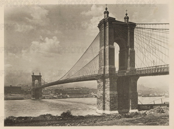 View of John A. Roebling Suspension Bridge at Cincinnati, The Ohio Suspension Bridge at Cincinnati, page, photomechanical print, Cincinnati, c. 1883 - in or before 1893, North America, paper, collotype, photoprint, height 140 mm x width 190 mm, suspension bridge, A sepia-toned photograph of a large suspension bridge spanning a river, showing a close-up of one massive stone tower with an arched opening and decorative finials on top, a network of vertical and diagonal suspension cables extending from the tower to the roadway, and the bridge deck with a latticework railing. The river flows beneath the bridge, with a vessel moored along the far bank and industrial buildings lining the waterfront beyond. The near riverbank in the foreground shows rough terrain and low vegetation, and the sky above contains scattered clouds.