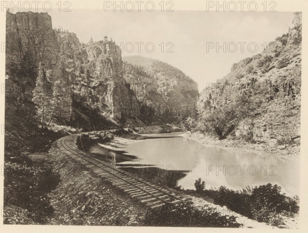 View of the Rio Grande with rock walls in Colorado, Echo Cliffs, Colorado - Denver & Rio Grande RR, page, photomechanical print, Colorado, c. 1883 - in or before 1893, North America, paper, collotype, photoprint, height 140 mm x width 191 mm, railway, train, river, inland cliff face, A sepia-toned landscape photograph showing a river running between steep rocky cliffs with trees scattered along the slopes and ridges. A curved set of railroad tracks follows the riverbank in the foreground, with wooden ties visible against a gravelly embankment. Reflections from the cliffs and vegetation appear on the calm surface of the water. The left side features a tall conifer near the base of a sheer rock face, while the right side shows terraced rock formations and scrubby vegetation. Distant hills rise behind the canyon walls beneath a pale, cloudless sky.