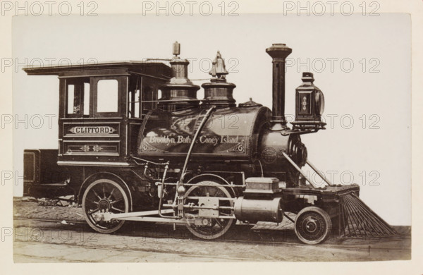 American steam locomotive, photograph, page, number: '53', United States of America, c. 1871 - in or before 1881, photographer, North America, paper, albumen print, height 124 mm x width 198 mm, Photo on page 125., steam locomotive, A sepia-toned photograph showing the side of a vintage steam locomotive with a rectangular cab featuring two windows and decorative paneling, a rounded boiler with ornate trim and raised cylindrical domes, a tall vertical smokestack, and a lantern mounted near the front. Visible mechanical linkages, pipes, and pistons run along the lower side above large spoked wheels and smaller front wheels, and a slatted metal pilot extends from the front near the rails. The locomotive rests on cobblestone pavement against a plain light background.