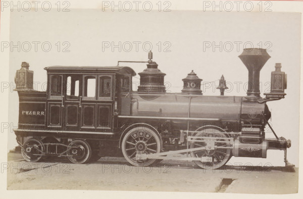 American steam locomotive, photograph, page, United States of America, c. 1871 - in or before 1881, photographer, North America, paper, albumen print, height 125 mm x width 200 mm, Photo on page 131., steam locomotive, A sepia-toned photograph of a steam locomotive shown in profile, occupying most of the frame. The engine has a rectangular cab with arched window openings and decorative paneling, a cylindrical boiler with domed fittings and a tall funnel, and visible driving wheels with patterned wheel centers and connecting rods. Ornate lamp housings or boxes are mounted at both ends of the cab and near the funnel, and a small bell sits atop the boiler. The locomotive rests on a ground surface with a small rectangular depression visible beneath the front wheel area. The image has a light background and a narrow border around the photograph.