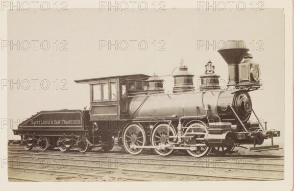 American steam locomotive, photograph, page, United States of America, c. 1871 - in or before 1881, photographer, North America, paper, albumen print, height 125 mm x width 199 mm, Photo on page 146., steam locomotive, A vintage steam locomotive and its tender sit on railroad tracks against a pale background, showing a large cylindrical boiler with domed fittings and piping, a tall flared smokestack, a square headlamp mounted at the front, and a round-faced front plate. The engineer's cab has framed windows and a projecting roof, and the tender behind it has a sloped top and visible wheel assemblies. Large driving wheels with spokes and connecting rods are visible beneath the boiler, along with exposed mechanical linkages and external piping. The scene is monochrome with subtle sepia tones, and the ground beneath the tracks is visible.