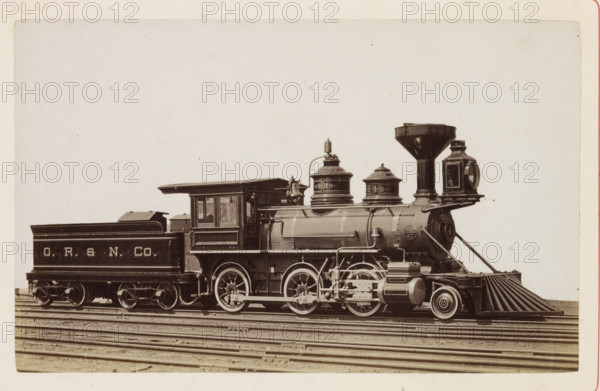 American freight steam locomotive, photograph, page, United States of America, c. 1871 - in or before 1881, photographer, North America, paper, albumen print, height 124 mm x width 198 mm, Photo on page 90., steam locomotive, A sepia-toned photograph of a vintage steam locomotive and its tender on parallel tracks, showing the side profile of the engine with a rounded smokebox, a tall flared smokestack, and two domed fittings on the boiler; the cab has rectangular windows and a roof overhang, and the tender sits behind with a sloped top. The visible running gear includes large spoked driving wheels, connecting rods, and various pipes and mechanical linkages along the side. At the front there is a metal pilot with slatted bars angling downward and a headlight housing mounted above the smokebox. The image shows the train centered horizontally against a pale background with the rails and sleepers beneath.