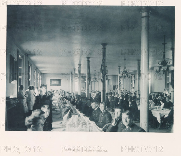 Cadet's mess-room, photograph, page, photographer: Edward H. Hart, United States Naval Academy, cliché maker: The Photo-gravure Co., New York (city), c. 1885 - in or before 1890, Edward H. Hart, The Photo-gravure Co., photographer, cliché maker, North America, Maryland, Annapolis, paper, collotype, photoprint, height 160 mm x width 215 mm, dining-room, officer, military academy, A large dining hall filled with long tables and many people seated along them, several uniformed individuals visible in the foreground, rows of standing columns running down the center of the room, chandeliers hanging from the ceiling, windows along the left wall, groups of staff in light-colored clothing standing along the left side, folded napkins and place settings on the tables, framed pictures on the walls, and flags hanging from some columns.