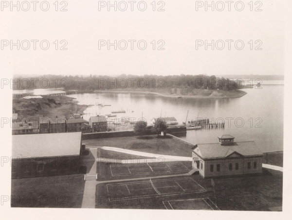 Looking north from tower, photograph, page, photographer: Edward H. Hart, United States Naval Academy, cliché maker: The Photo-gravure Co., New York (city), c. 1885 - in or before 1890, North America, Maryland, Annapolis, paper, collotype, photoprint, height 167 mm x width 238 mm, gulf, bay, A sepia-toned aerial view shows a waterfront scene with calm water, a curved shoreline lined with trees, and a small peninsula with a grassy slope. Several piers and docks extend into the water with small sailboats and rowboats moored nearby. A row of low buildings with pitched roofs sits along the near shore, and a larger building with a cupola and a facade facing the water occupies the foreground beside paved walkways and marked rectangular courts. Paths and grassy areas surround the buildings, and a distant treeline and a long bridge are visible along the horizon.