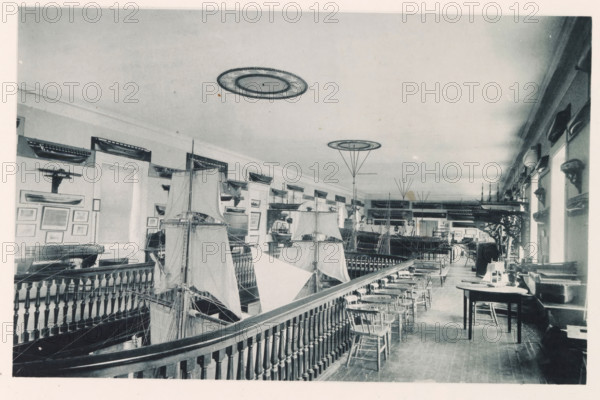 Seamanship-model-room, photograph, page, photographer: Edward H. Hart, United States Naval Academy, cliché maker: The Photo-gravure Co., New York (city), c. 1885 - in or before 1890, North America, Maryland, Annapolis, paper, collotype, photoprint, height 150 mm x width 238 mm, ship-model, interior, representation of a building, A long interior room photographed from an elevated railing view lined with turned wooden balusters and topped by a row of wooden chairs, filled with large model sailing ships with tall masts and billowed sails occupying the floor and display stands, walls adorned with mounted hull models and framed pictures, a series of windows along the right wall letting in light, tables and workbenches with tools and smaller models on the right side, decorative circular ceiling fixtures, and various wooden shelves and display cases extending along the length of the room.
