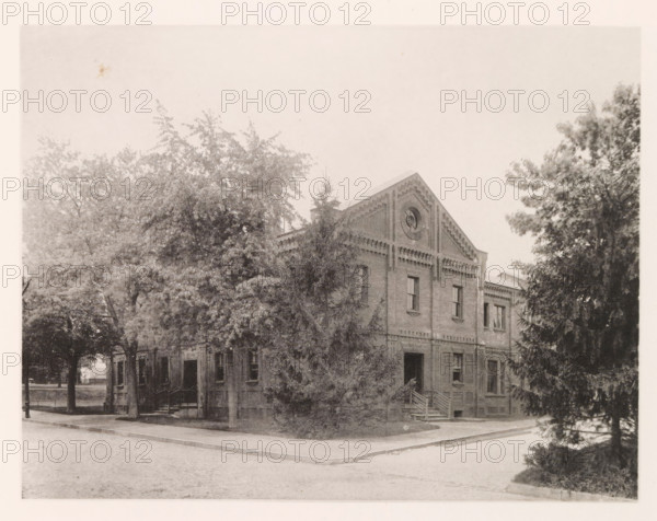Physical laboratory, photograph, page, photographer: Edward H. Hart, United States Naval Academy, cliché maker: The Photo-gravure Co., New York (city), c. 1885 - in or before 1890, North America, Maryland, Annapolis, paper, collotype, photoprint, height 177 mm x width 231 mm, laboratory, research room, military academy, A sepia-toned photograph shows a brick building with a triangular gable featuring a circular decorative opening; the facade has rectangular windows and a set of exterior steps leading to a doorway. Large trees with dense foliage partially obscure the building on both sides and in front, and a paved street runs in the foreground with a curb and sidewalk along the building. The scene has a slightly faded, aged appearance and soft contrast throughout.