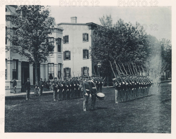 Military parade on the grounds of the United States Naval Academy in Annapolis, photograph, page, photographer: Edward H. Hart, United States Naval Academy, cliché maker: The Photo-gravure Co., New York (city), c. 1885 - in or before 1890, North America, Maryland, Annapolis, paper, collotype, photoprint, height 177 mm x width 236 mm, military parade, pageant, military academy, A wide lawn in front of a multi-story brick building with tall windows and shutters is bordered by mature trees, and a long line of uniformed soldiers stands at attention in formation holding rifles with bayonets, several men in front carrying a drum and wearing caps, while a few other figures stand near the building entrance and under the trees.